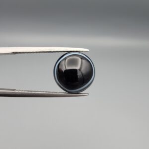 Multi-angle macro photographs of a 5.97-carat natural African Banded Eye Agate (Sulemani Aqeeq) held in gem tweezers, showing a top view with a dominant black body framed by a sharp white arc, a profile view with a distinct V-shaped white fortification band, and a reverse view revealing a multi-eye cellular pattern in blue-grey and black.