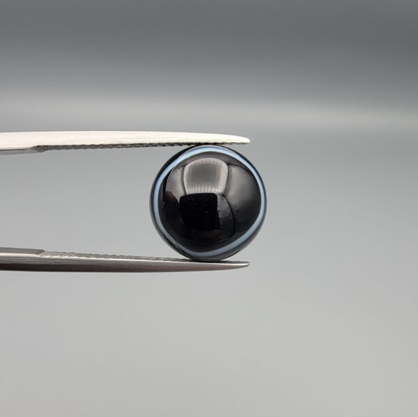 Multi-angle macro photographs of a 5.97-carat natural African Banded Eye Agate (Sulemani Aqeeq) held in gem tweezers, showing a top view with a dominant black body framed by a sharp white arc, a profile view with a distinct V-shaped white fortification band, and a reverse view revealing a multi-eye cellular pattern in blue-grey and black.