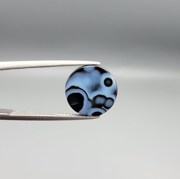Multi-angle macro photographs of a 5.97-carat natural African Banded Eye Agate (Sulemani Aqeeq) held in gem tweezers, showing a top view with a dominant black body framed by a sharp white arc, a profile view with a distinct V-shaped white fortification band, and a reverse view revealing a multi-eye cellular pattern in blue-grey and black.