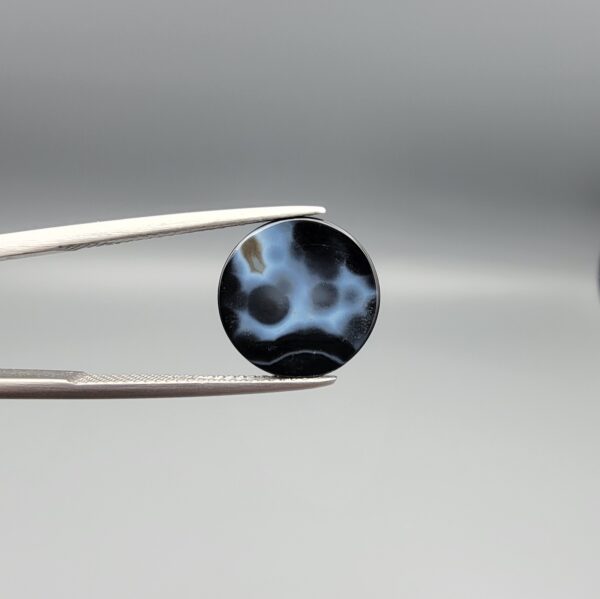 Multi-angle macro photographs of a 12.13-carat natural African Banded Eye Agate (Sulemani Aqeeq) held in steel gem tweezers, showcasing a top view with concentric white and dark brown fortification bands surrounding a translucent core with a dark eye inclusion, a profile view displaying layered fortification banding, and a reverse view revealing multiple rounded eye motifs and cellular banding against a dark chalcedony field.