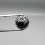 Multi-angle macro photographs of a 12.13-carat natural African Banded Eye Agate (Sulemani Aqeeq) held in steel gem tweezers, showcasing a top view with a mirror-polished deep black body bordered by a fine white ring, a side profile displaying lower white flank banding, and a reverse view revealing a unique luminous blue-grey eye motif on a flat black field.