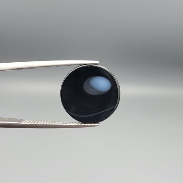 Multi-angle macro photographs of a 12.13-carat natural African Banded Eye Agate (Sulemani Aqeeq) held in steel gem tweezers, showcasing a top view with a mirror-polished deep black body bordered by a fine white ring, a side profile displaying lower white flank banding, and a reverse view revealing a unique luminous blue-grey eye motif on a flat black field.