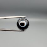 Multi-angle macro photographs of a 6.29-carat natural African Banded Eye Agate (Sulemani Aqeeq) held in steel gem tweezers, showcasing a top view with a central black pupil enclosed by a crisp white halo ring, a profile view displaying lower flank fortification bands, and a reverse view revealing a flat translucent milky-grey gradient base.