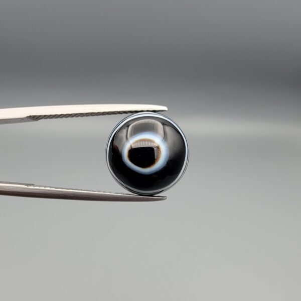 Multi-angle macro photographs of a 6.29-carat natural African Banded Eye Agate (Sulemani Aqeeq) held in steel gem tweezers, showcasing a top view with a central black pupil enclosed by a crisp white halo ring, a profile view displaying lower flank fortification bands, and a reverse view revealing a flat translucent milky-grey gradient base.