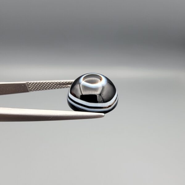 Multi-angle macro photographs of a 6.29-carat natural African Banded Eye Agate (Sulemani Aqeeq) held in steel gem tweezers, showcasing a top view with a central black pupil enclosed by a crisp white halo ring, a profile view displaying lower flank fortification bands, and a reverse view revealing a flat translucent milky-grey gradient base.