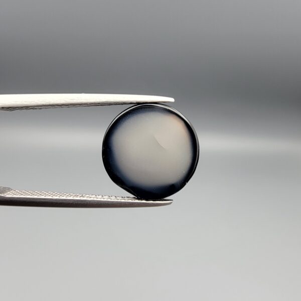 Multi-angle macro photographs of a 6.29-carat natural African Banded Eye Agate (Sulemani Aqeeq) held in steel gem tweezers, showcasing a top view with a central black pupil enclosed by a crisp white halo ring, a profile view displaying lower flank fortification bands, and a reverse view revealing a flat translucent milky-grey gradient base.
