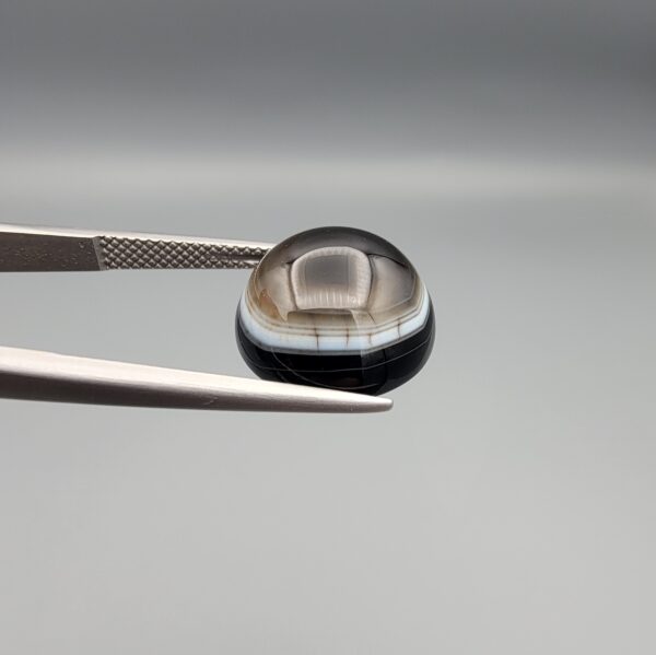 Multi-angle macro photographs of a 9.53-carat natural African Banded Eye Agate (Sulemani Aqeeq) held in steel gem tweezers, showcasing a top view with a translucent brownish-grey central field framed by a sharp concentric ring, a side profile revealing layered fortification banding along the flanks, and a smooth jet-black flat reverse base.