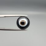 Multi-angle macro photographs of a 6.20-carat natural African Banded Eye Agate (Sulemani Aqeeq) held in steel gem tweezers, displaying a top view with a deep black core enclosed by a sharp concentric white ring, a side profile revealing smooth high-dome curvature, and a reverse view featuring a flat, solid black base with subtle natural mineral matrix markings.
