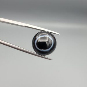 Multi-angle macro photographs of a 12.17-carat natural African Banded Eye Agate (Sulemani Aqeeq) held in steel gem tweezers, showcasing a top view with a sharp white concentric halo ring enclosing an intense black core, a side profile displaying smooth high-dome geometry with flank banding, and a reverse view of a flat nebulous dark chalcedony base.