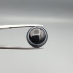 Multi-angle macro photographs of an 8.57-carat natural African Banded Eye Agate (Sulemani Aqeeq) held in steel gem tweezers, showcasing a top view with a crisp concentric outer white ring framing a deep black core, a side profile displaying high-dome geometry and layered fortification pinstripes, and a reverse view of a flat, solid mirror-polished black base.
