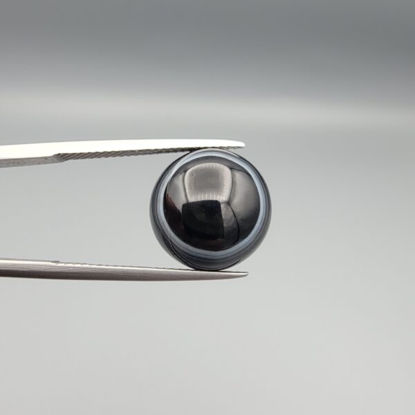 Multi-angle macro photographs of an 8.57-carat natural African Banded Eye Agate (Sulemani Aqeeq) held in steel gem tweezers, showcasing a top view with a crisp concentric outer white ring framing a deep black core, a side profile displaying high-dome geometry and layered fortification pinstripes, and a reverse view of a flat, solid mirror-polished black base.
