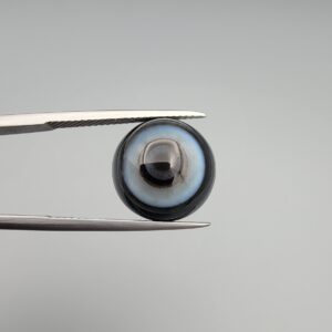 Multi-angle macro photographs of an 8.35-carat natural African Banded Eye Agate (Sulemani Aqeeq) held in steel gem tweezers, showcasing a top view with concentric black, grey, and white rings framing a luminous core, a side profile displaying smooth high-dome geometry with layered fortification bands, and a reverse view of a flat, dark mottled base with a nebulous center.