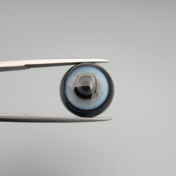 Multi-angle macro photographs of an 8.35-carat natural African Banded Eye Agate (Sulemani Aqeeq) held in steel gem tweezers, showcasing a top view with concentric black, grey, and white rings framing a luminous core, a side profile displaying smooth high-dome geometry with layered fortification bands, and a reverse view of a flat, dark mottled base with a nebulous center.