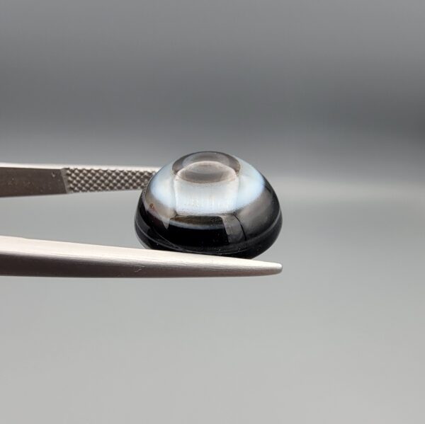 Multi-angle macro photographs of an 8.35-carat natural African Banded Eye Agate (Sulemani Aqeeq) held in steel gem tweezers, showcasing a top view with concentric black, grey, and white rings framing a luminous core, a side profile displaying smooth high-dome geometry with layered fortification bands, and a reverse view of a flat, dark mottled base with a nebulous center.