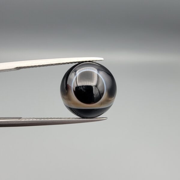 Multi-angle macro photographs of a 7.64-carat natural African Banded Eye Agate (Sulemani Aqeeq) held in steel gem tweezers, showcasing a top view with a sharp concentric ring and lower translucent band, a side profile displaying smooth high-dome geometry with horizontal flank fortification, and a reverse view of a flat black base featuring a striking horizontal banded division.