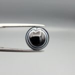 Multi-angle macro photographs of a 6.57-carat natural African Banded Eye Agate (Sulemani Aqeeq) held in steel gem tweezers, showcasing a top view with a rare heart-shaped concentric fortification ring pattern, a side profile revealing smooth high-dome geometry with layered fortification banding, and a flat, solid black reverse base with natural micro-inclusions.