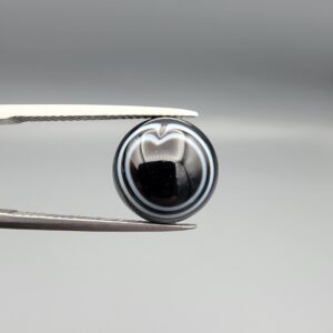 Multi-angle macro photographs of a 6.57-carat natural African Banded Eye Agate (Sulemani Aqeeq) held in steel gem tweezers, showcasing a top view with a rare heart-shaped concentric fortification ring pattern, a side profile revealing smooth high-dome geometry with layered fortification banding, and a flat, solid black reverse base with natural micro-inclusions.