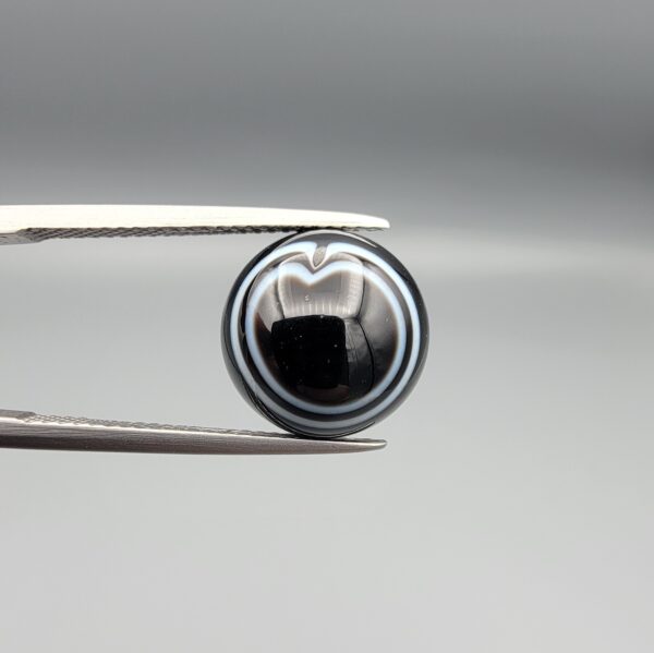 Multi-angle macro photographs of a 6.57-carat natural African Banded Eye Agate (Sulemani Aqeeq) held in steel gem tweezers, showcasing a top view with a rare heart-shaped concentric fortification ring pattern, a side profile revealing smooth high-dome geometry with layered fortification banding, and a flat, solid black reverse base with natural micro-inclusions.