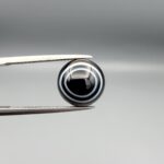 Multi-angle macro photographs of a 5.89-carat natural African Banded Eye Agate (Sulemani Aqeeq) held in steel gem tweezers, displaying a top view with a crisp concentric white ring framing a dark brownish-black central core, and a reverse view revealing a flat, dark mottled chalcedony base with subtle natural edge inclusions.