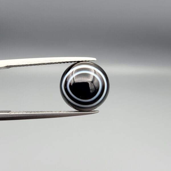 Multi-angle macro photographs of a 5.89-carat natural African Banded Eye Agate (Sulemani Aqeeq) held in steel gem tweezers, displaying a top view with a crisp concentric white ring framing a dark brownish-black central core, and a reverse view revealing a flat, dark mottled chalcedony base with subtle natural edge inclusions.
