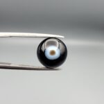 Multi-angle macro photographs of a 5.62-carat natural African Banded Eye Agate (Sulemani Aqeeq) held in steel gem tweezers, showcasing a top view with a central brown pupil core encircled by a white halo ring set within a deep black body, a side profile displaying smooth high-dome geometry with lower flank fortification, and a reverse view revealing a flat translucent milky-blue base with a solid black eclipse patch.