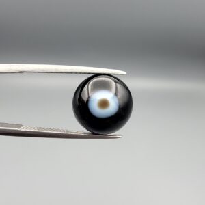 Multi-angle macro photographs of a 5.62-carat natural African Banded Eye Agate (Sulemani Aqeeq) held in steel gem tweezers, showcasing a top view with a central brown pupil core encircled by a white halo ring set within a deep black body, a side profile displaying smooth high-dome geometry with lower flank fortification, and a reverse view revealing a flat translucent milky-blue base with a solid black eclipse patch.