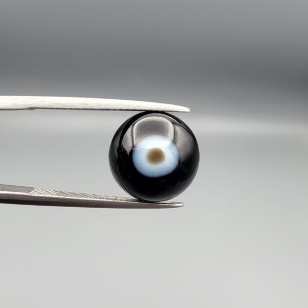 Multi-angle macro photographs of a 5.62-carat natural African Banded Eye Agate (Sulemani Aqeeq) held in steel gem tweezers, showcasing a top view with a central brown pupil core encircled by a white halo ring set within a deep black body, a side profile displaying smooth high-dome geometry with lower flank fortification, and a reverse view revealing a flat translucent milky-blue base with a solid black eclipse patch.