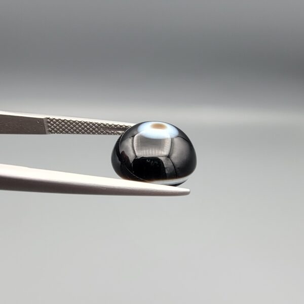 Multi-angle macro photographs of a 5.62-carat natural African Banded Eye Agate (Sulemani Aqeeq) held in steel gem tweezers, showcasing a top view with a central brown pupil core encircled by a white halo ring set within a deep black body, a side profile displaying smooth high-dome geometry with lower flank fortification, and a reverse view revealing a flat translucent milky-blue base with a solid black eclipse patch.