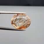 High-resolution macro photograph of an 8.63-carat natural Rutilated Quartz (Moh-e-Najaf) held in steel tweezers, showing a finely faceted oval cut, water-clear crystal clarity, and a dense internal lattice of golden-brown and bronze rutile needles sourced from Najaf Al-Ashraf, Iraq.