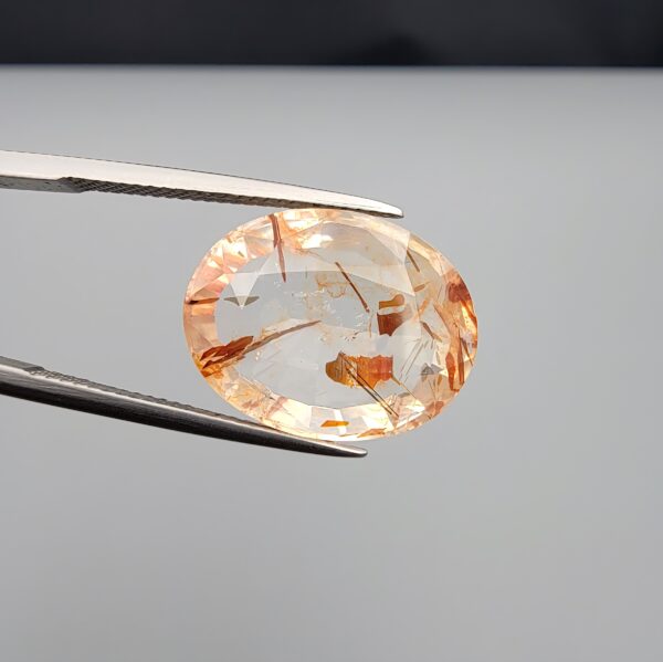 High-resolution macro photographs of a 14.26-carat natural Rutilated Quartz (Moh-e-Najaf) held in steel tweezers, displaying a brilliant oval cut, exceptional water-clear crystal matrix, and natural internal reddish-orange hematite platelets with fine scattered rutile needles sourced from Najaf Al-Ashraf, Iraq.