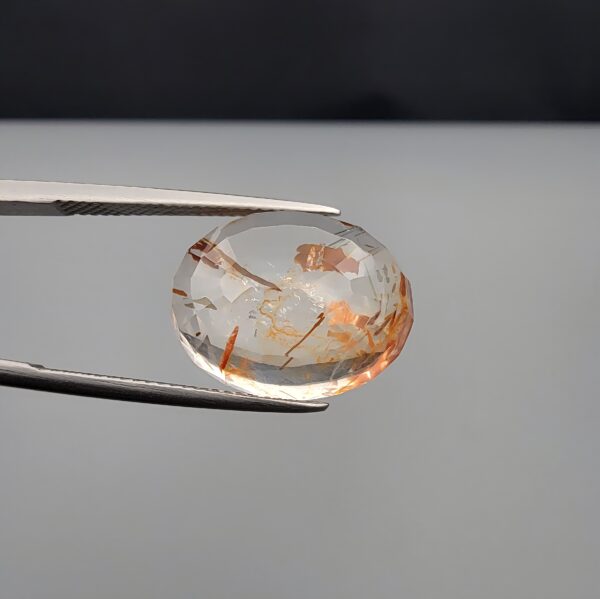 High-resolution macro photographs of a 14.26-carat natural Rutilated Quartz (Moh-e-Najaf) held in steel tweezers, displaying a brilliant oval cut, exceptional water-clear crystal matrix, and natural internal reddish-orange hematite platelets with fine scattered rutile needles sourced from Najaf Al-Ashraf, Iraq.