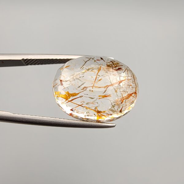High-resolution macro photographs of a 17.27-carat natural Rutilated Quartz (Moh-e-Najaf) held in steel tweezers, displaying a finely faceted oval silhouette, water-clear crystal body, and rich internal golden-brown hematite platelets with contrasting dark rutile needles sourced from Najaf Al-Ashraf, Iraq.
