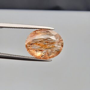 High-resolution macro photograph of an 8.45-carat natural Rutilated Quartz (Moh-e-Najaf) held in steel gem tweezers, showcasing a brilliant oval facet cut, high crystal transparency, and a dense, horizontally aligned internal lattice of golden-red and dark rutile needles sourced from Najaf Al-Ashraf, Iraq.