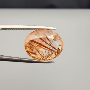 High-resolution macro photographs of an 8.99-carat natural Rutilated Quartz (Moh-e-Najaf) held in steel tweezers, showcasing a masterfully faceted oval silhouette, water-clear quartz body, and striking diagonal reddish-brown rutile blade inclusions sourced from Najaf Al-Ashraf, Iraq.