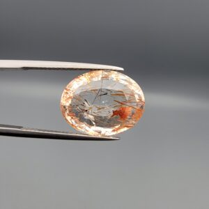 High-resolution macro photographs of a 14.39-carat natural Rutilated Quartz (Moh-e-Najaf) held in steel gem tweezers, displaying a brilliantly faceted oval cut, water-clear crystal body, fine golden rutile needles, and a focal concentrated orange-red hematite inclusion cluster sourced from Najaf Al-Ashraf, Iraq.