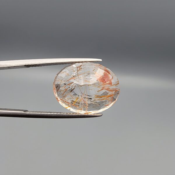 High-resolution macro photographs of a 14.39-carat natural Rutilated Quartz (Moh-e-Najaf) held in steel gem tweezers, displaying a brilliantly faceted oval cut, water-clear crystal body, fine golden rutile needles, and a focal concentrated orange-red hematite inclusion cluster sourced from Najaf Al-Ashraf, Iraq.