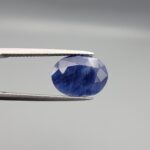 High-resolution macro photograph of a 5.38-carat natural Afghan Blue Indigo Sapphire (Neelam) held in steel tweezers, showcasing a finely faceted oval cut, deep indigo-blue color saturation, and natural corundum silk inclusions sourced from Afghanistan.