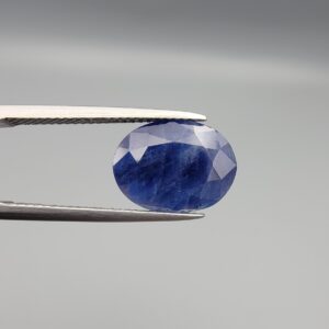 High-resolution macro photograph of a 5.38-carat natural Afghan Blue Indigo Sapphire (Neelam) held in steel tweezers, showcasing a finely faceted oval cut, deep indigo-blue color saturation, and natural corundum silk inclusions sourced from Afghanistan.