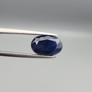 High-resolution macro photograph of a 5.11-carat natural Afghan Blue Indigo Sapphire (Neelam) held in steel gem tweezers, showcasing an oval facet cut, deep midnight indigo-blue body color, and natural corundum silk texture sourced from Afghanistan.