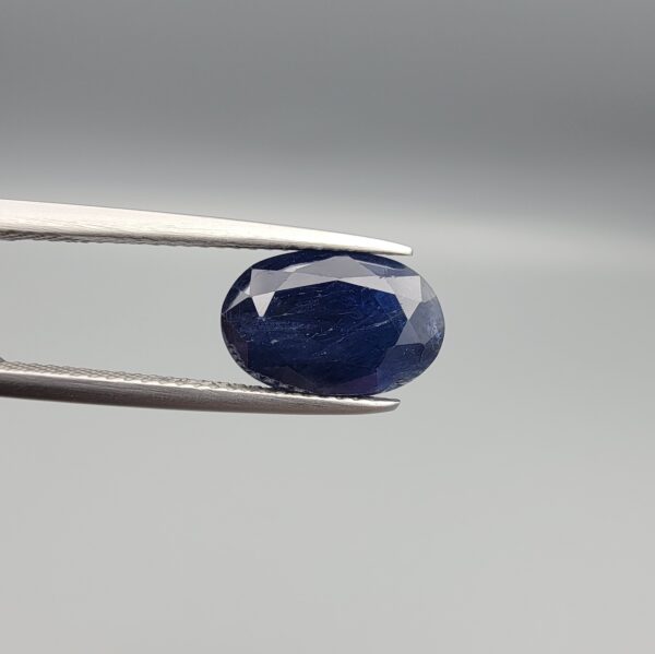 High-resolution macro photograph of a 5.11-carat natural Afghan Blue Indigo Sapphire (Neelam) held in steel gem tweezers, showcasing an oval facet cut, deep midnight indigo-blue body color, and natural corundum silk texture sourced from Afghanistan.