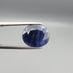 High-resolution macro photographs of a 10.26-carat natural Afghan Blue Indigo Sapphire (Neelam) held in steel tweezers, showcasing a masterfully faceted oval cut, rich midnight indigo-blue body color, and natural internal corundum silk structures sourced from Afghanistan.