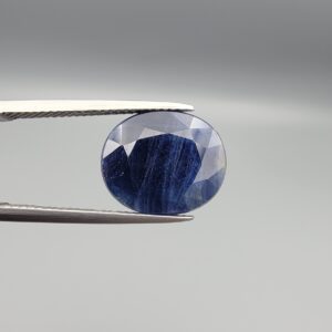 High-resolution macro photographs of a 10.26-carat natural Afghan Blue Indigo Sapphire (Neelam) held in steel tweezers, showcasing a masterfully faceted oval cut, rich midnight indigo-blue body color, and natural internal corundum silk structures sourced from Afghanistan.