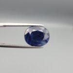 High-resolution macro photographs of a 9.87-carat natural Afghan Blue Indigo Sapphire (Neelam) held in steel tweezers, showcasing a brilliant oval facet cut, deep indigo-blue color saturation, and natural internal corundum silk features sourced from Afghanistan.