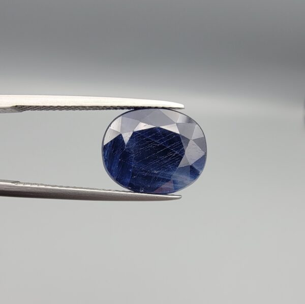 High-resolution macro photographs of a 9.87-carat natural Afghan Blue Indigo Sapphire (Neelam) held in steel tweezers, showcasing a brilliant oval facet cut, deep indigo-blue color saturation, and natural internal corundum silk features sourced from Afghanistan.