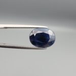 High-resolution macro photographs of a 6.70-carat natural Afghan Blue Indigo Sapphire (Neelam) held in steel gem tweezers, showcasing a brilliant oval facet cut, deep indigo-blue color saturation, and natural internal corundum silk structures sourced from Afghanistan.