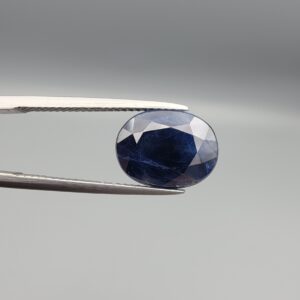 High-resolution macro photographs of a 6.70-carat natural Afghan Blue Indigo Sapphire (Neelam) held in steel gem tweezers, showcasing a brilliant oval facet cut, deep indigo-blue color saturation, and natural internal corundum silk structures sourced from Afghanistan.