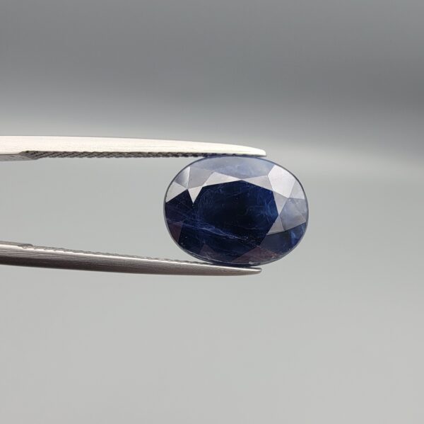 High-resolution macro photographs of a 6.70-carat natural Afghan Blue Indigo Sapphire (Neelam) held in steel gem tweezers, showcasing a brilliant oval facet cut, deep indigo-blue color saturation, and natural internal corundum silk structures sourced from Afghanistan.