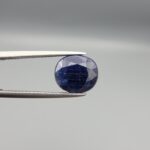 High-resolution macro photographs of a 5.40-carat natural Afghan Blue Indigo Sapphire (Neelam) held in steel gem tweezers, showcasing a masterfully faceted oval silhouette, deep midnight indigo-blue body color, and natural internal corundum silk structures sourced from Afghanistan.