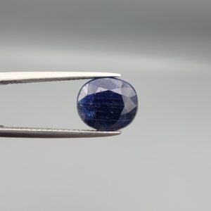 High-resolution macro photographs of a 5.40-carat natural Afghan Blue Indigo Sapphire (Neelam) held in steel gem tweezers, showcasing a masterfully faceted oval silhouette, deep midnight indigo-blue body color, and natural internal corundum silk structures sourced from Afghanistan.