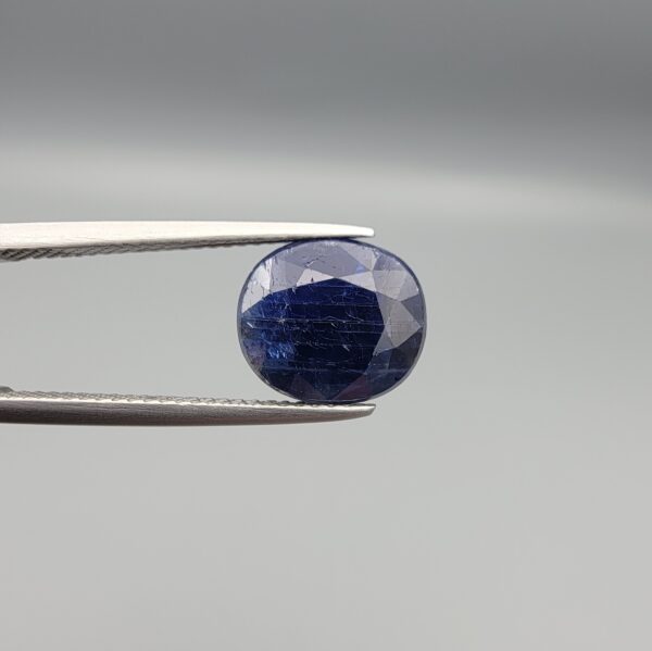 High-resolution macro photographs of a 5.40-carat natural Afghan Blue Indigo Sapphire (Neelam) held in steel gem tweezers, showcasing a masterfully faceted oval silhouette, deep midnight indigo-blue body color, and natural internal corundum silk structures sourced from Afghanistan.